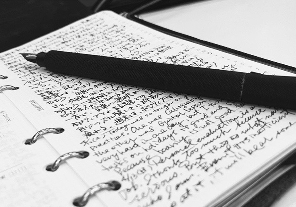 A black and white image of a black pen laying across a filled page of a PLOTTER leather binder.