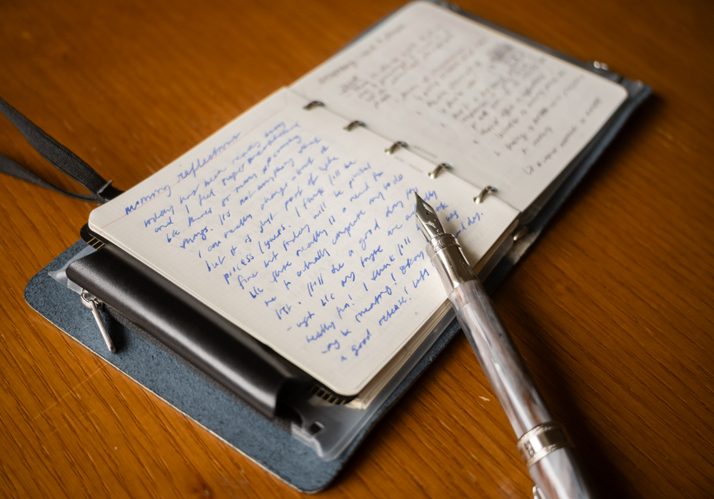 A PLOTTER Leather Binder in Mini 5 Square Size open to a page full of morning journaling beside a fountain pen.