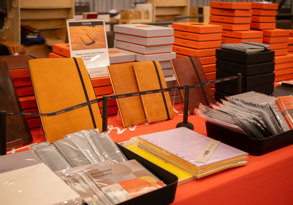 A person shopping at the PLOTTER USA table filled with PLOTTER items, leather binders, and stationery at the California Pen Show.