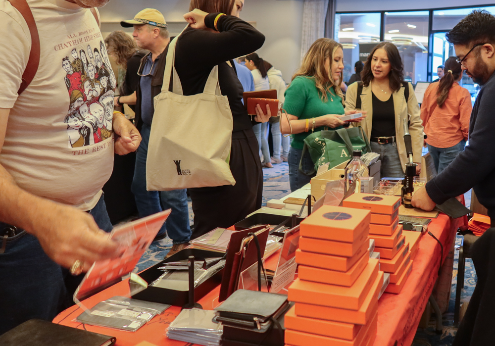 Customers shopping at the PLOTTER USA table filled with PLOTTER items, leather binders, and stationery at the California Pen Show.