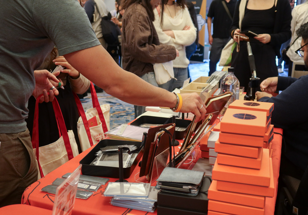 A person shopping at the PLOTTER USA table filled with PLOTTER items, leather binders, and stationery at the California Pen Show.
