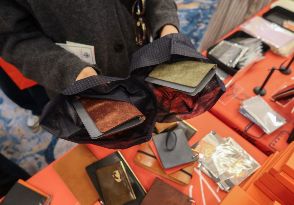 A person shopping at the PLOTTER USA table, trying to decide between two different colors (Brown or Olive) of the Mini 5 Pueblo Leather Binders.