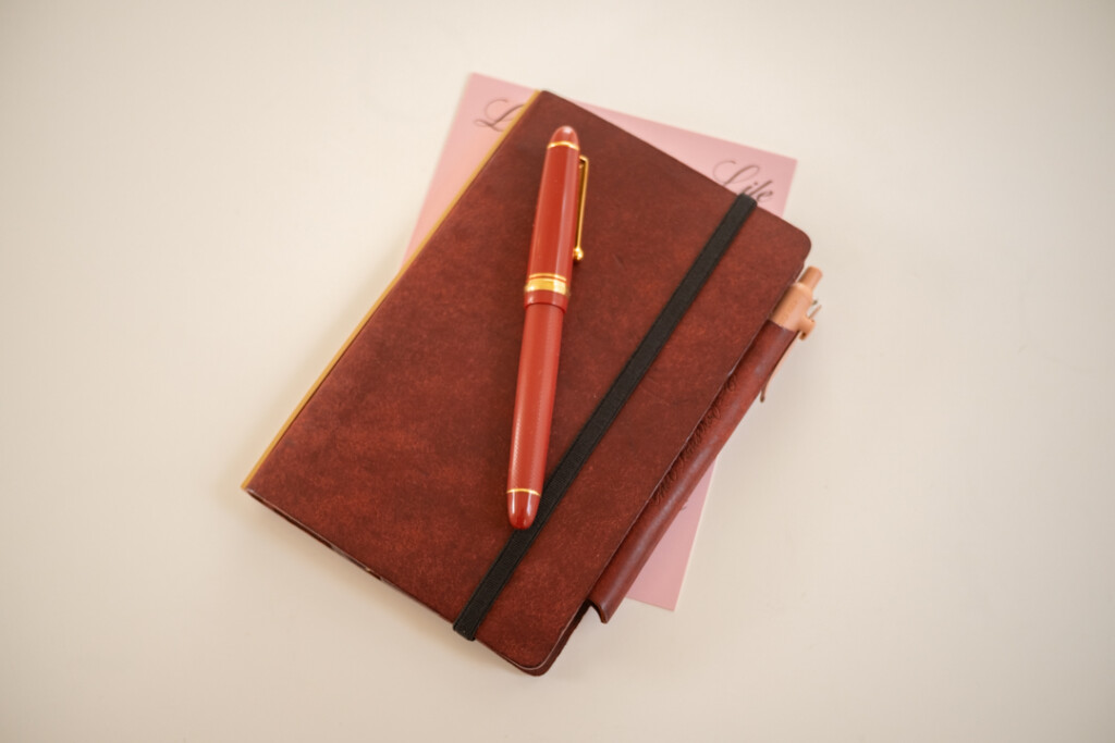 A Red Brown PLOTTER Leather Binder in Bible Size, with a red fountain pen on top of it on a white table.