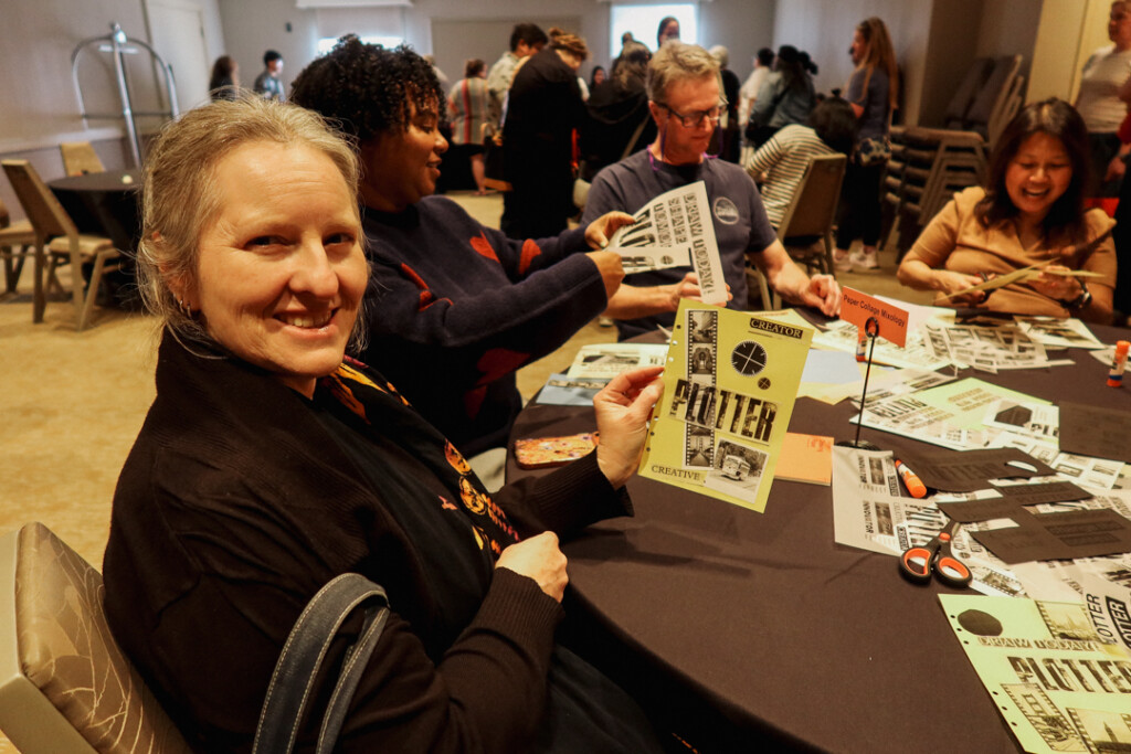 A person showing off their finished collages at a collaging station on a round table filled with paper supplies and glue.
