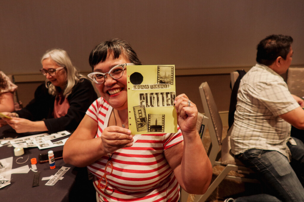 A person showing off their finished collages at a collaging station on a round table filled with paper supplies and glue.