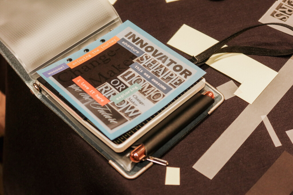 A person showing off their finished collages at a collaging station on a round table filled with paper supplies and glue.