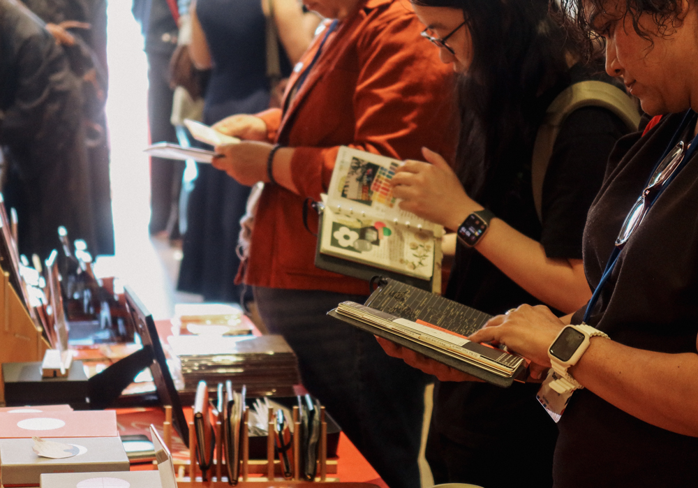 A person shopping at the PLOTTER USA table filled with PLOTTER items, leather binders, and stationery at the San Francisco Stationery Fest.