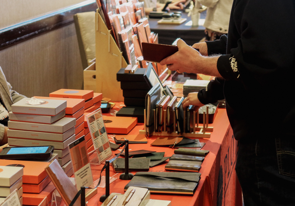 A person shopping at the PLOTTER USA table filled with PLOTTER items, leather binders, and stationery at the San Francisco Stationery Fest.