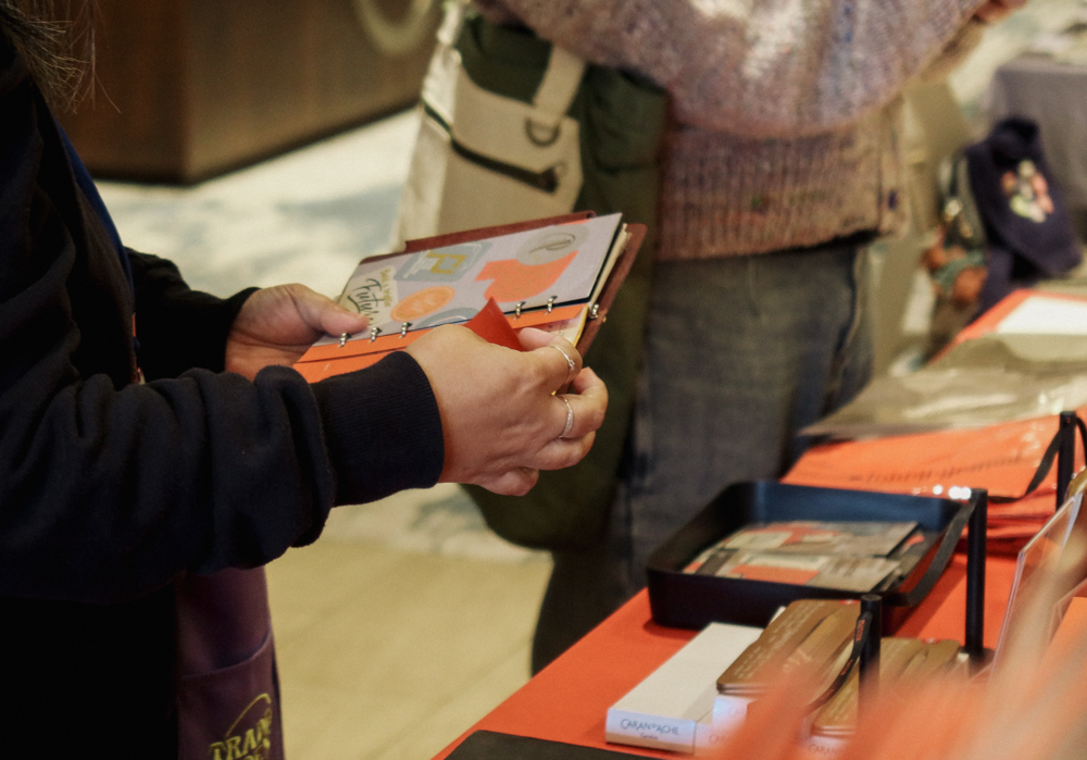 A person shopping at the PLOTTER USA table filled with PLOTTER items, leather binders, and stationery at the San Francisco Stationery Fest.