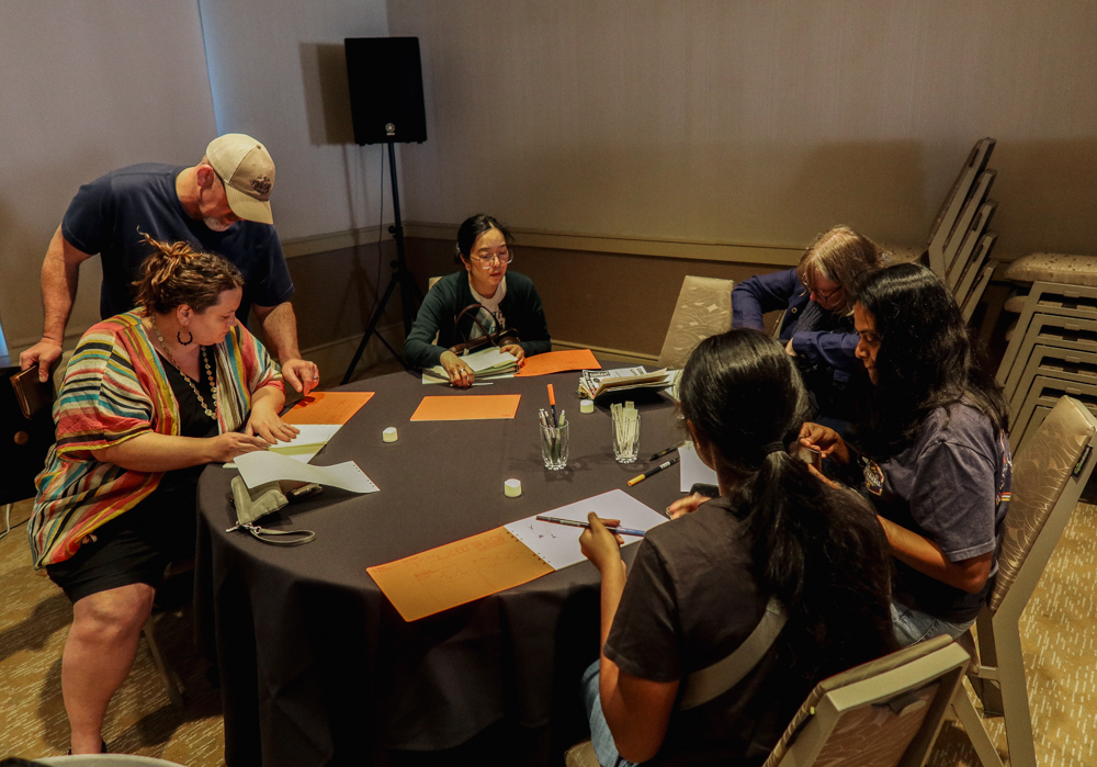 People are working on finishing their collages at a collaging station on a round table filled with paper supplies and glue.