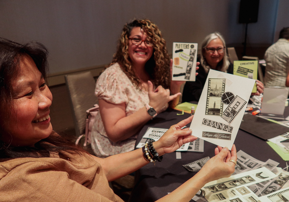 3 people showing off their finished collages at a collaging station on a round table filled with paper supplies and glue.