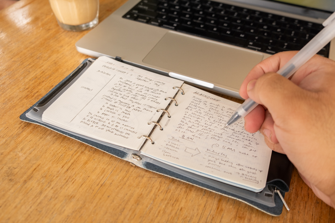 A PLOTTER Leather Binder in Mini 5 Square size opened on a wooden table beside a laptop, a cup of coffee, and a person writing in the binder.