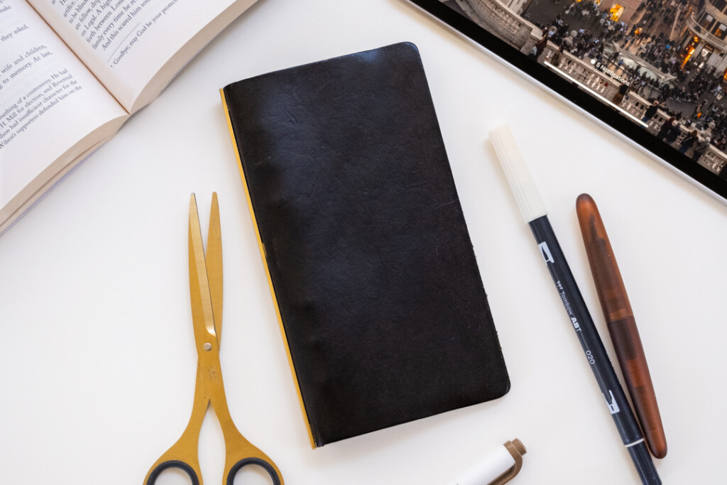 A PLOTTER Leather Binder in Black Pueblo Leather beside desk tools, a book, and a tablet.