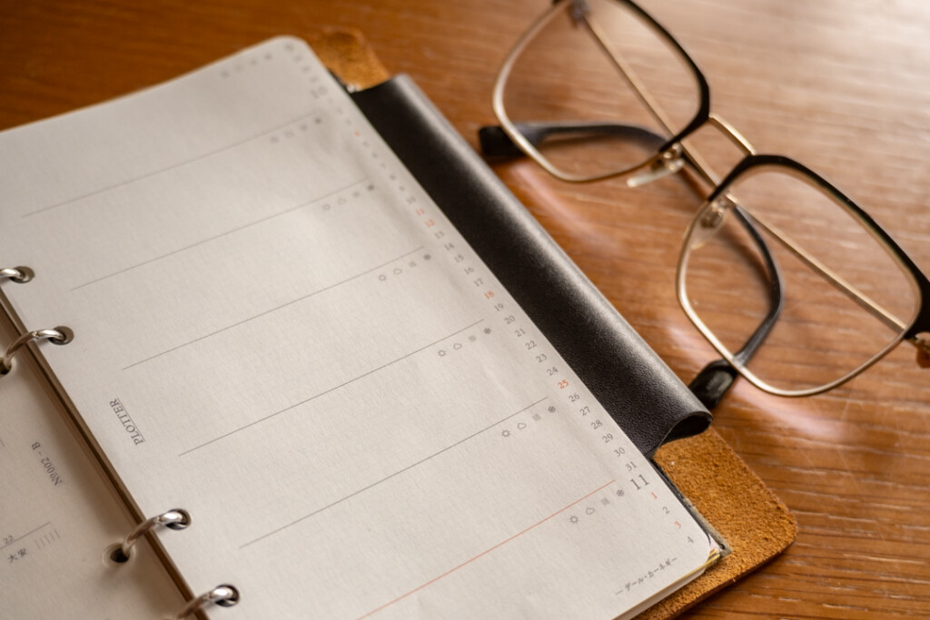 A close-up of a blank PLOTTER Weekly Schedule in a PLOTTER Leather Binder on a wooden desk which is beside reading glasses.