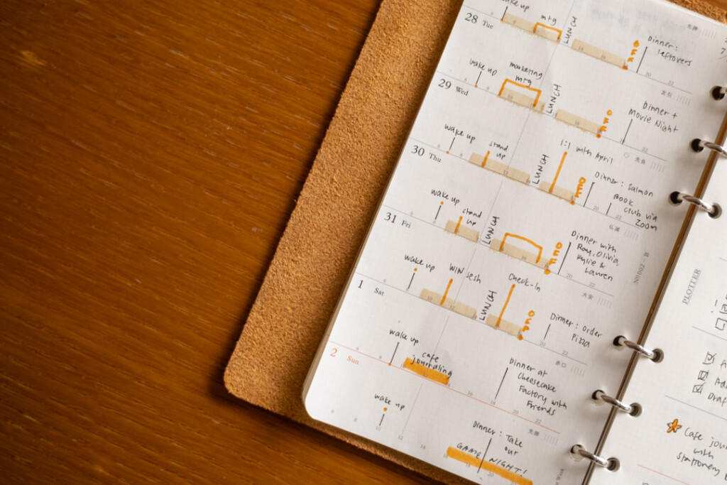 A filled-in PLOTTER Weekly Schedule showing the timeline side of the spread which is in a PLOTTER Leather Binder on a wooden desk.