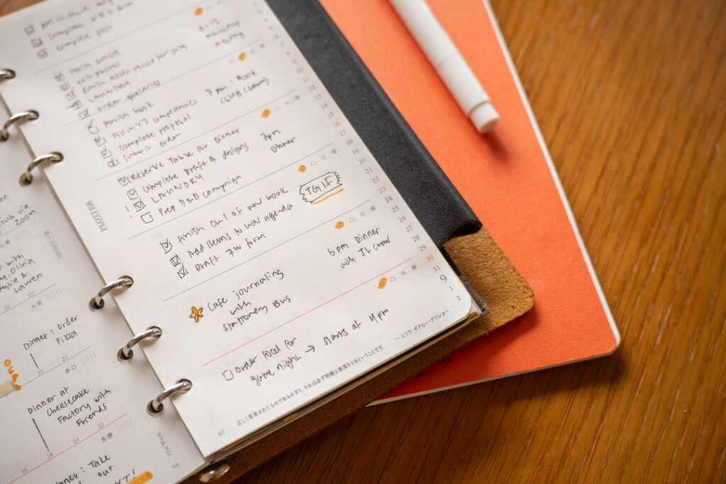 A filled-in PLOTTER Weekly Schedule showing the memo side of the spread which is in a PLOTTER Leather Binder on a wooden desk surrounded by writing utensil and scissors.