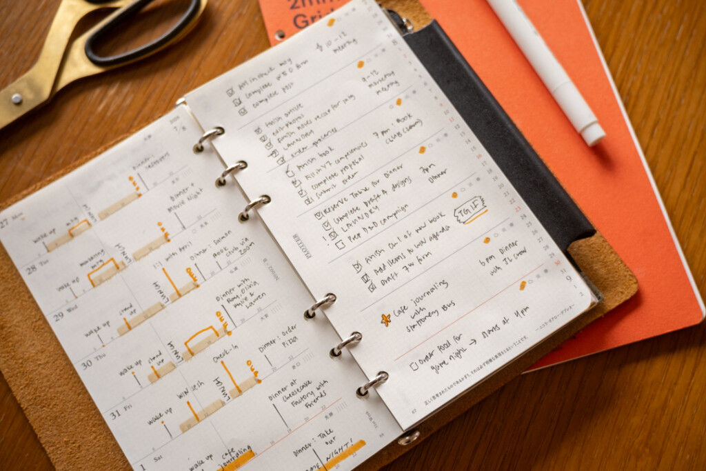 A filled-in PLOTTER Weekly Schedule in a PLOTTER Leather Binder on a wooden desk surrounded by writing utensils, paper pad and scissors.