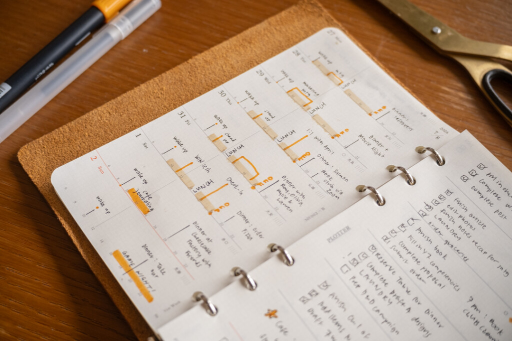 A filled-in PLOTTER Weekly Schedule showing the timeline side of the spread which is in a PLOTTER Leather Binder on a wooden desk surrounded by writing utensil and scissors.