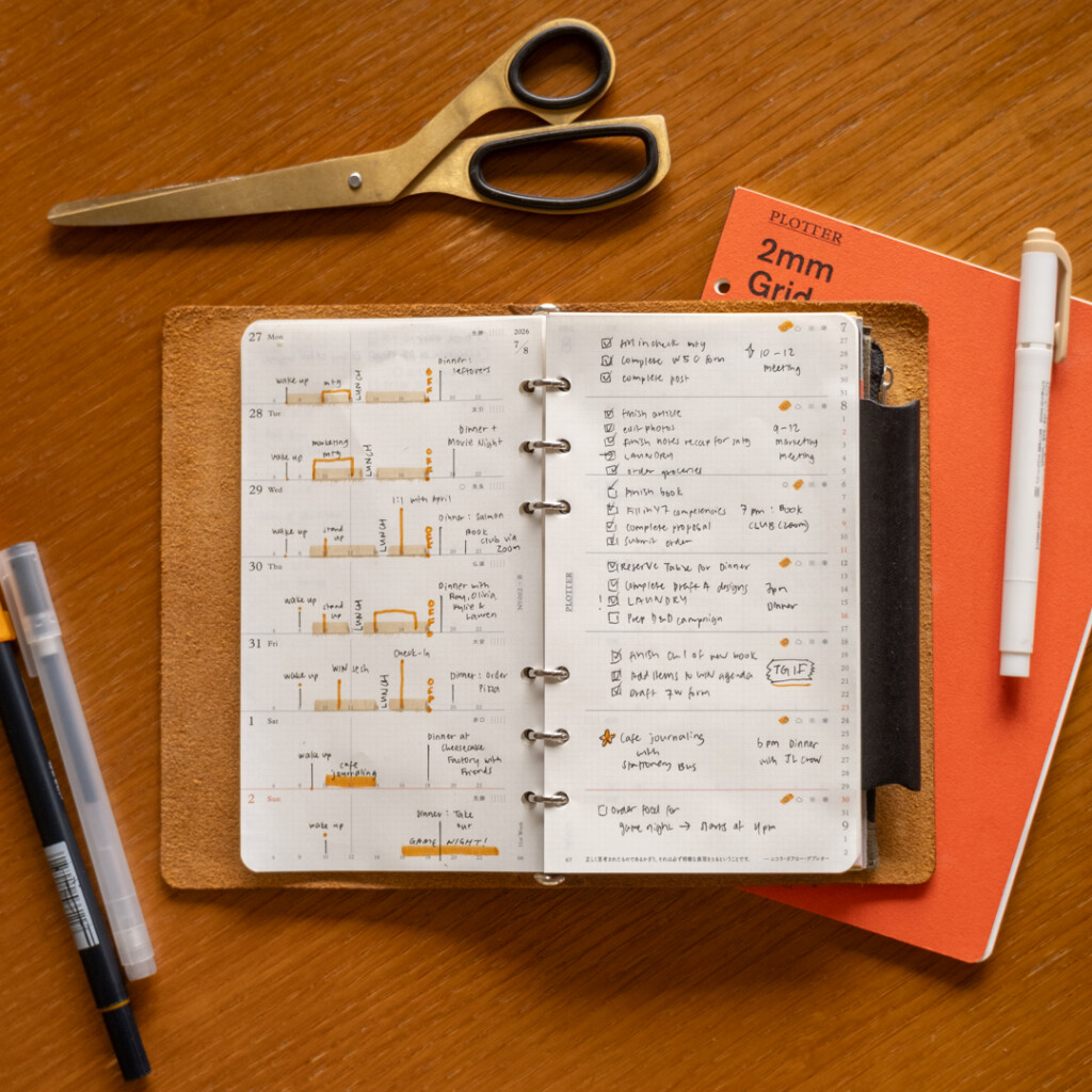A filled-in PLOTTER Weekly Schedule in a PLOTTER Leather Binder on a wooden desk surrounded by writing utensils, paper pad and scissors.