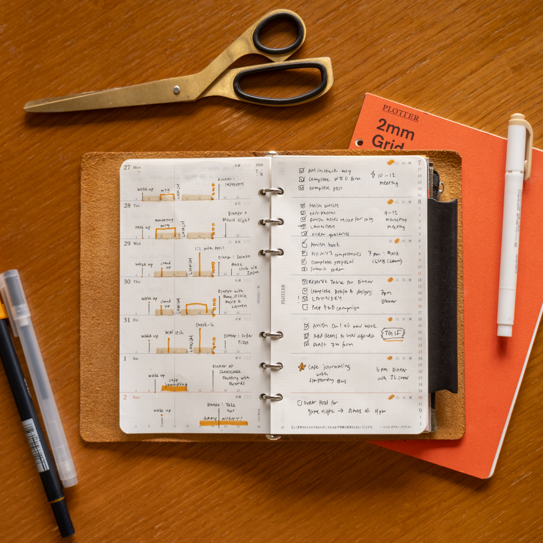 A filled-in PLOTTER Weekly Schedule in a PLOTTER Leather Binder on a wooden desk surrounded by writing utensils, paper pad and scissors.