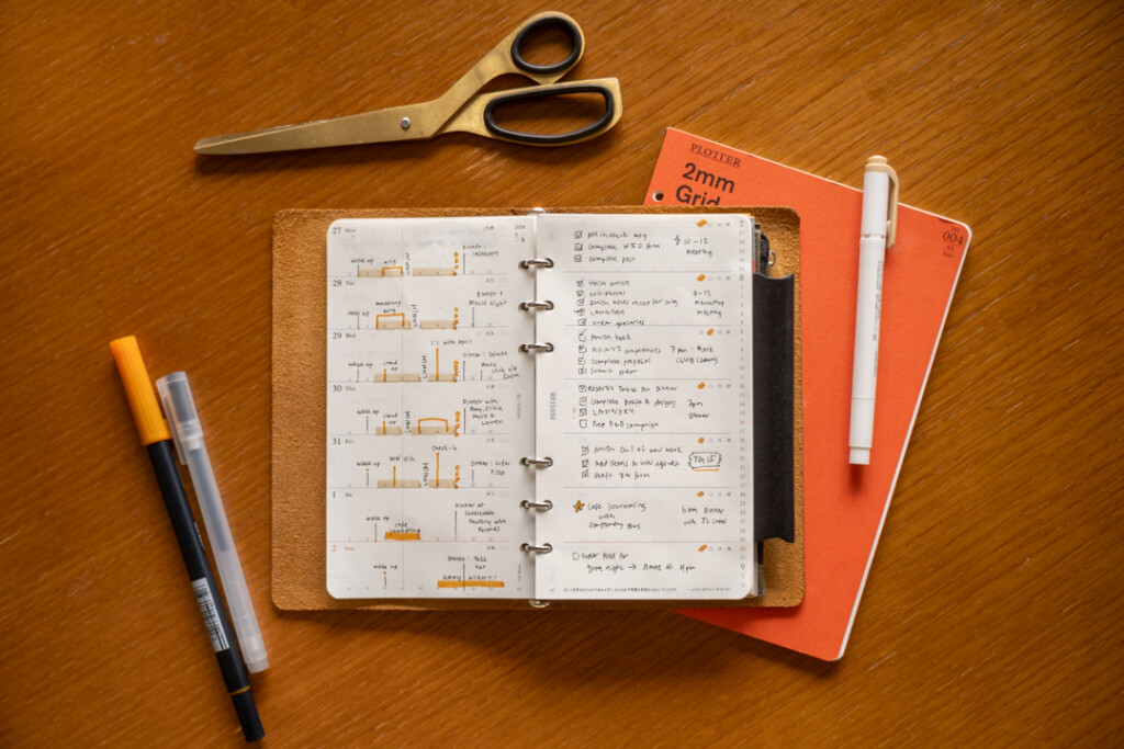 A filled-in PLOTTER Weekly Schedule in a PLOTTER Leather Binder on a wooden desk surrounded by writing utensils, paper pad and scissors.