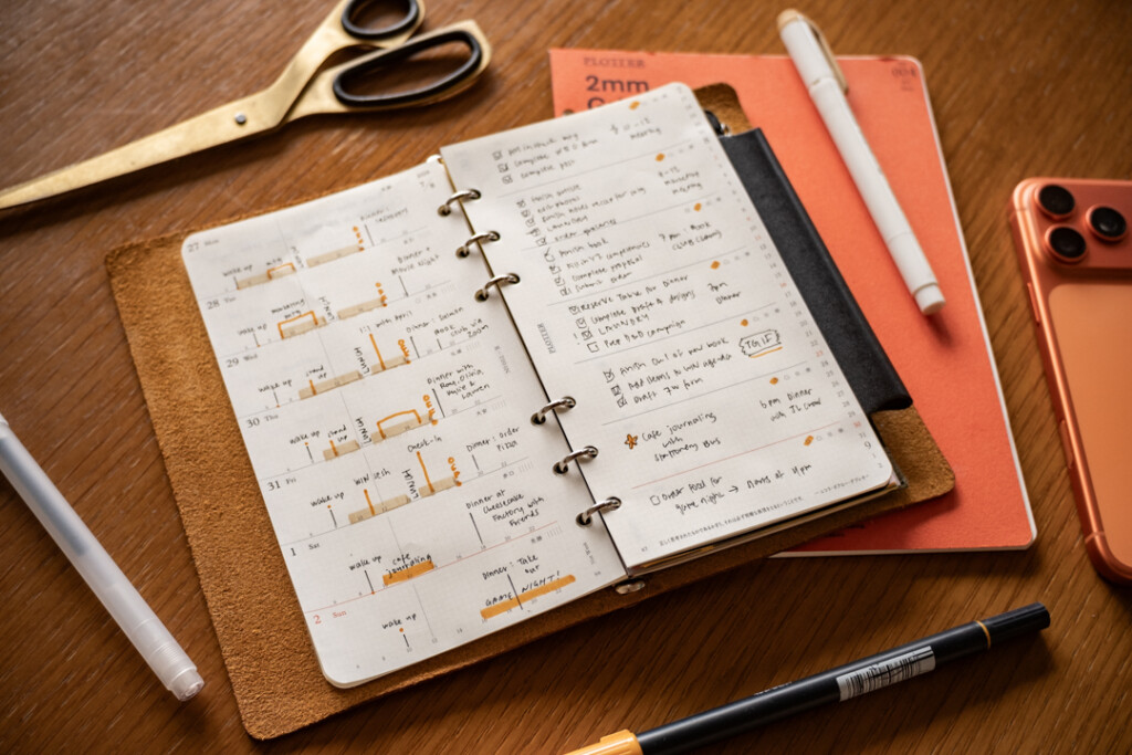 A filled-in PLOTTER Weekly Schedule in a PLOTTER Leather Binder on a wooden desk surrounded by writing utensils, paper pad and scissors.