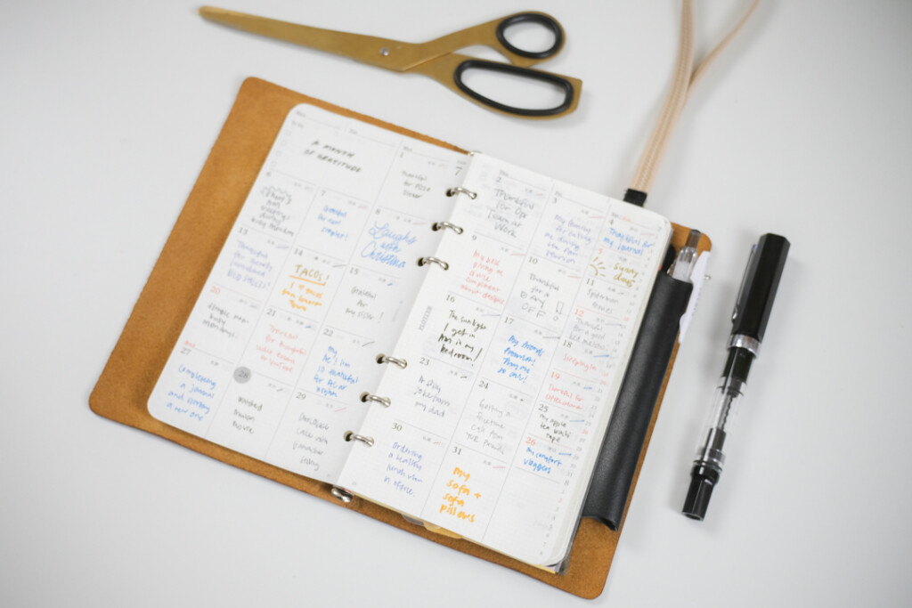 A PLOTTER Leather Binder is open to a monthly schedule page, which is filled in with different color pens as a daily gratitude log. There is a pen and scissors beside the leather binder.