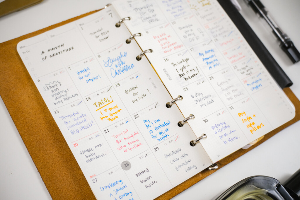 A PLOTTER Leather Binder is open to a monthly schedule page, which is filled in with different color pens as a daily gratitude log.