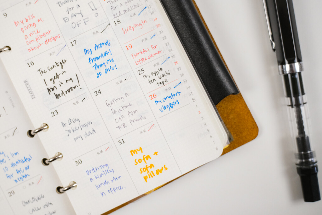 A close-up of a PLOTTER Leather Binder opened to a monthly schedule page, which is filled in with different color pens as a daily gratitude log.
