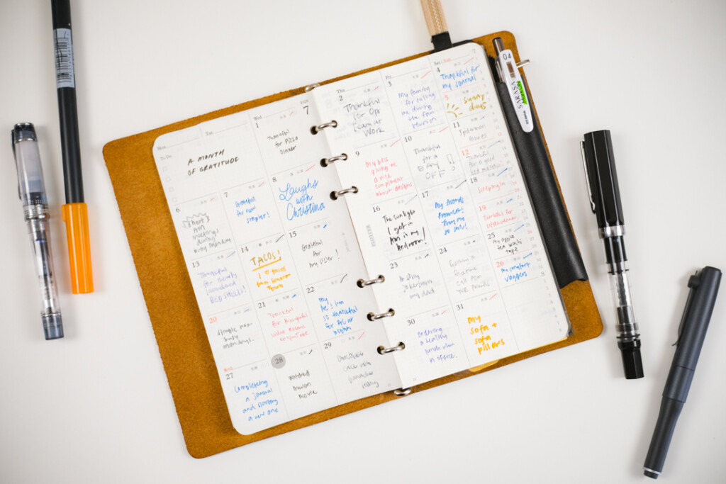 A PLOTTER Leather Binder is open to a monthly schedule page, which is filled in with different color pens as a daily gratitude log. There are different pens and markers beside the open leather binder.